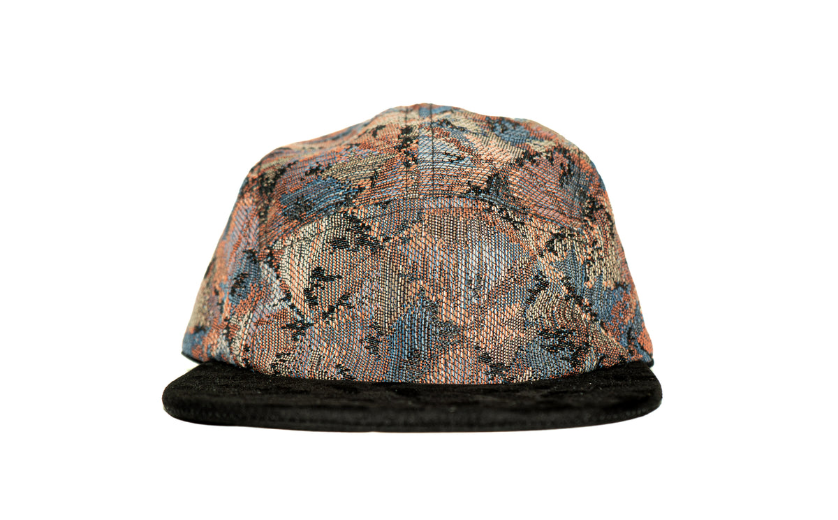 A structured five-panel cap featuring a textured, woven geometric pattern in shades of blue, orange, beige, and black, with a solid black flat brim.
